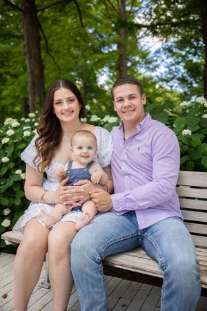 A family sits together on a wooden bench outdoors surrounded by lush green foliage and white flowering bushes.