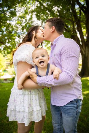 A happy baby in blue overalls smiles while being held outdoors in a sunny park with green trees in the background.
