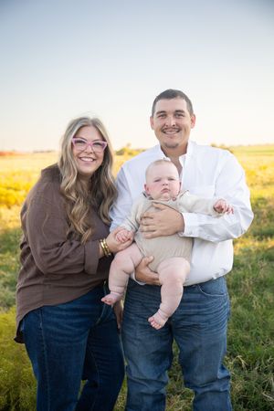 A family poses together in a golden field at sunset, with a small baby wearing light colored clothing.