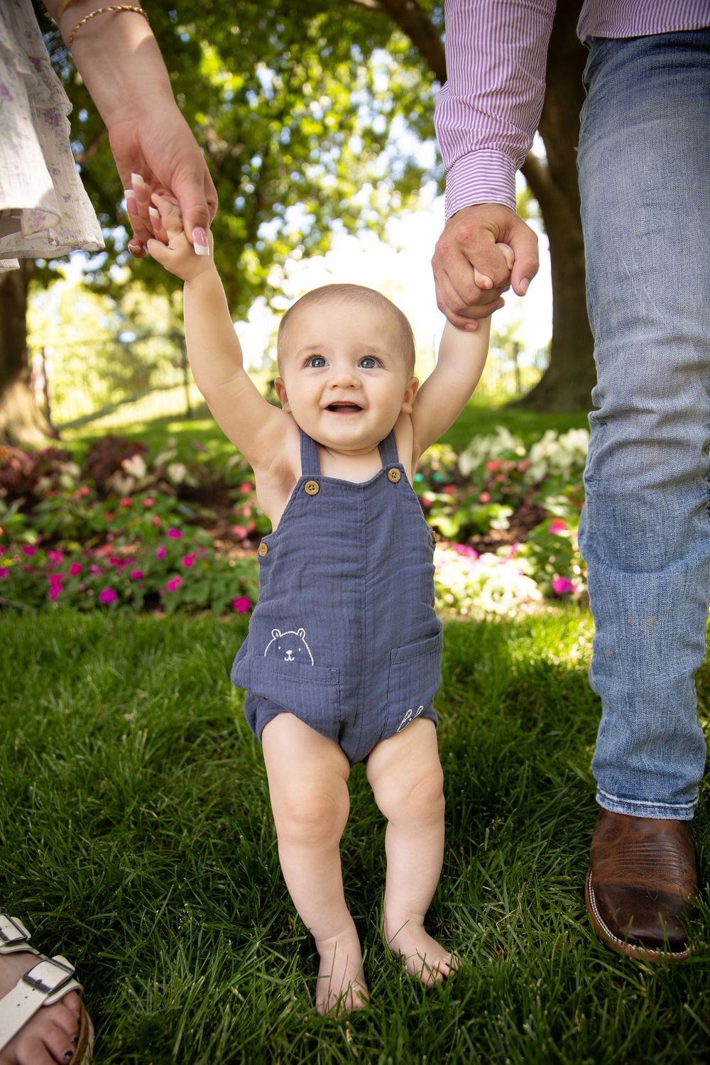 A baby in denim overalls holds hands with adults while learning to walk on green grass in a sunny garden setting.