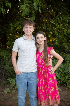 Two young people pose outdoors in casual attire - one in a patterned polo shirt and jeans, one in a bright pink floral dress.