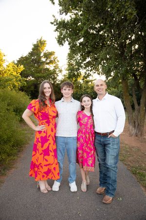 A family of four poses together outdoors wearing bright pink dresses and white shirts on a tree-lined path at sunset.