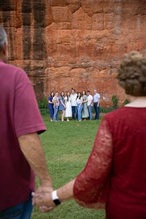 A group of people in formal attire pose together against a rustic red brick wall with grass in the foreground.