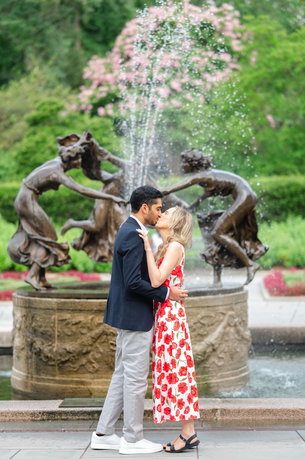 A couple shares a kiss after they get engaged in the Central Park Conservatory Garden in front of an ornate fountain with bronze sculptures.