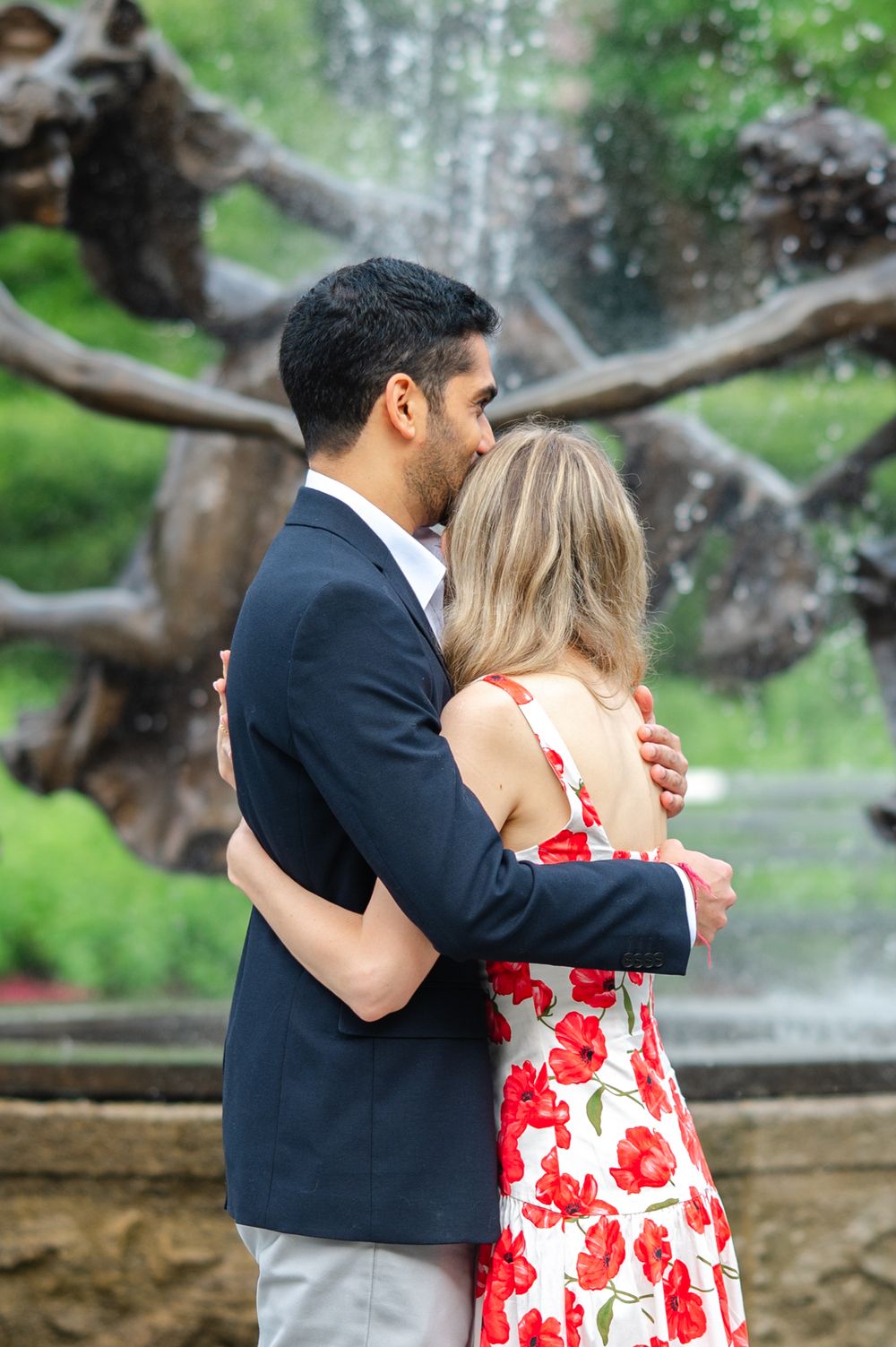 Couple embraces in a romantic moment outdoors with a red floral dress and navy suit against a natural backdrop.