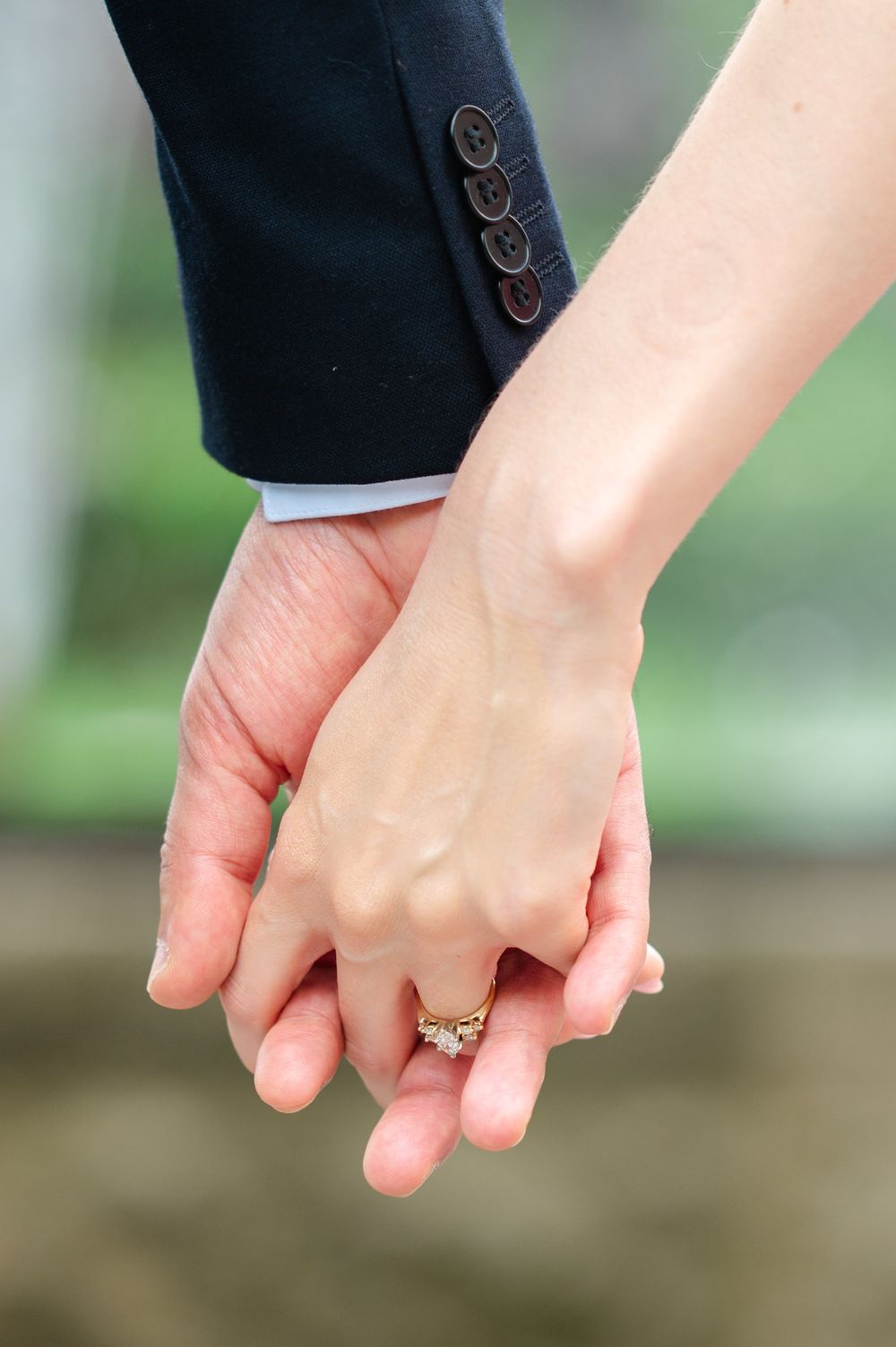 Detailed view of hands holding with a sparkling engagement ring in focus against a blurred garden background.