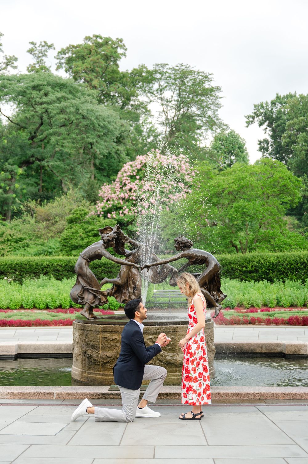 A couple shares a romantic moment in the Central Park Conservatory Garden setting with a decorative fountain in the background.