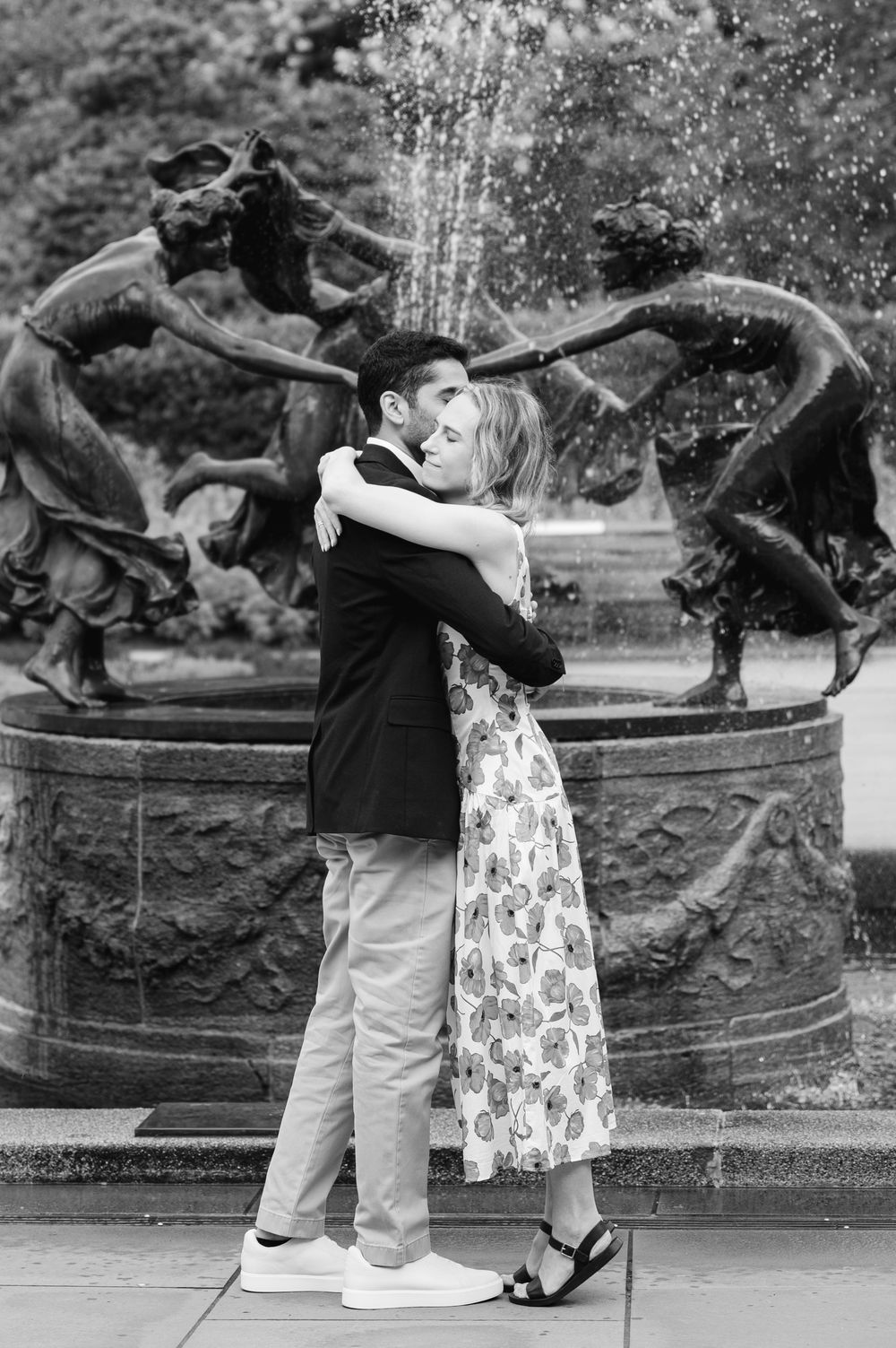 A couple hugging captured in black and white by a decorative fountain with dancing figure sculptures during their proposal.