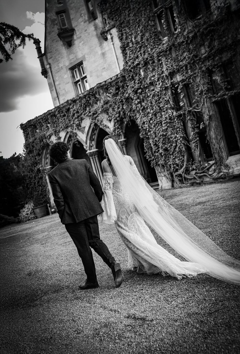 A black and white wedding photograph showing a couple walking together on a gravel path beside an ivy-covered building.