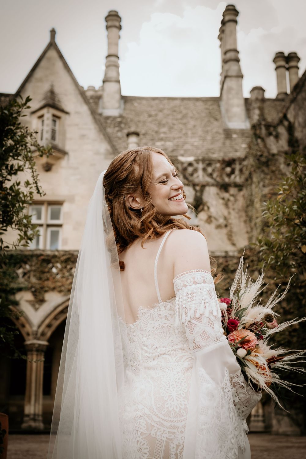 Elegant bride in lace off-shoulder dress poses gracefully against historic stone building backdrop.