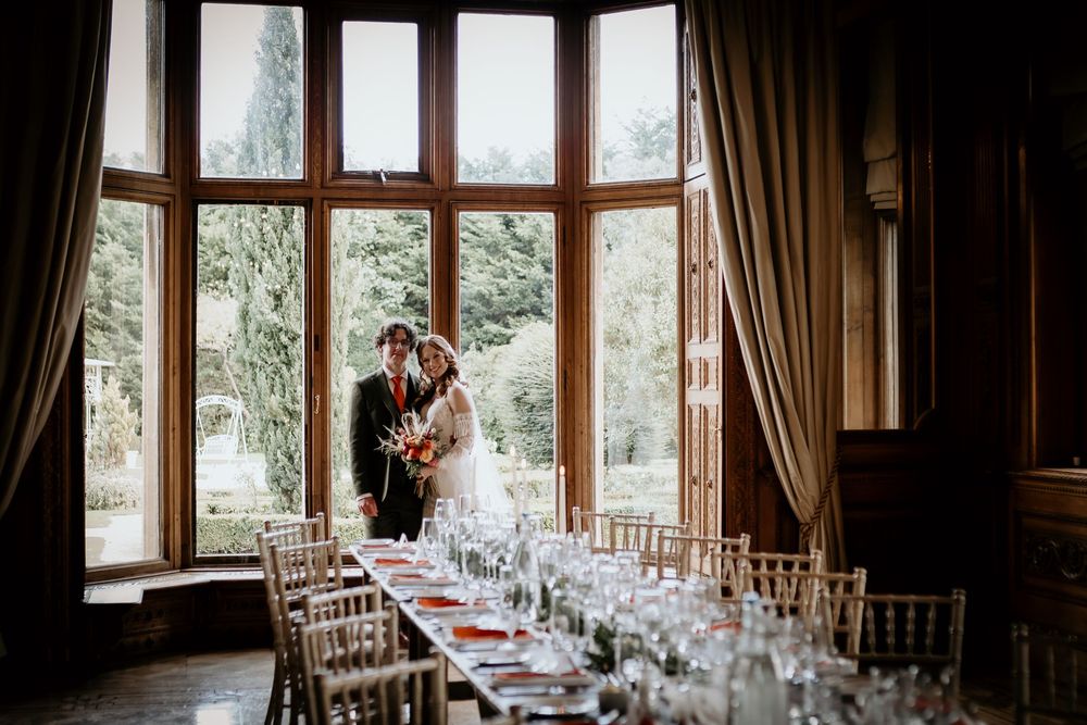Wedding reception tables set with glassware and floral centerpieces in an elegant wood-paneled room with large windows.