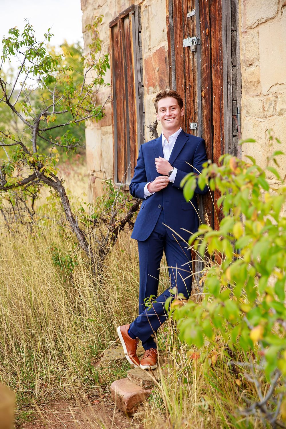 A series of outdoor portraits showing someone in a navy suit posing against a rustic stone wall with greenery.