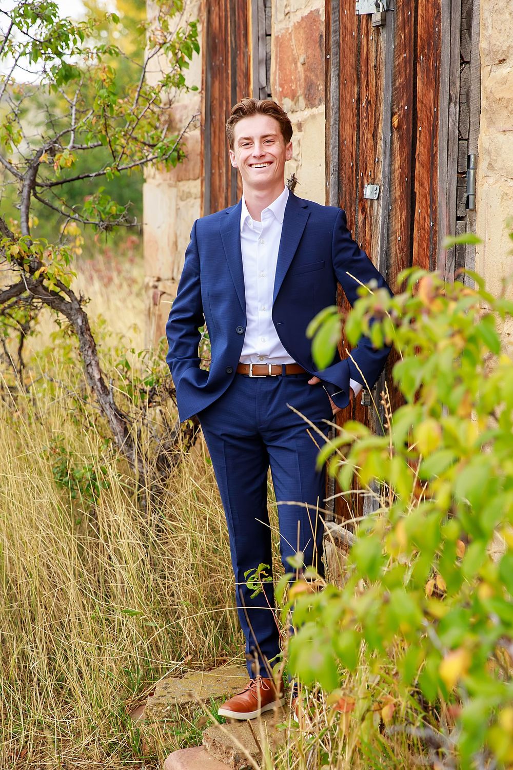 Professional portrait of someone in a navy suit and brown shoes standing against a rustic wall with greenery.