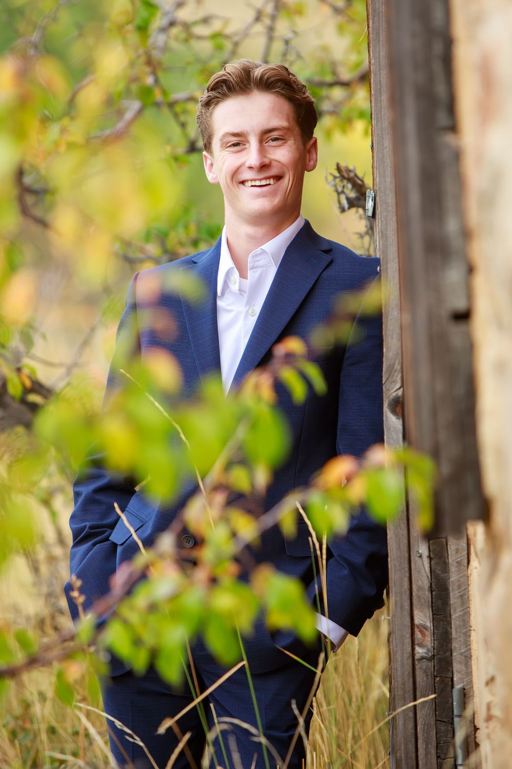 A person in a navy suit poses casually against a wooden fence with blurred greenery in the background.