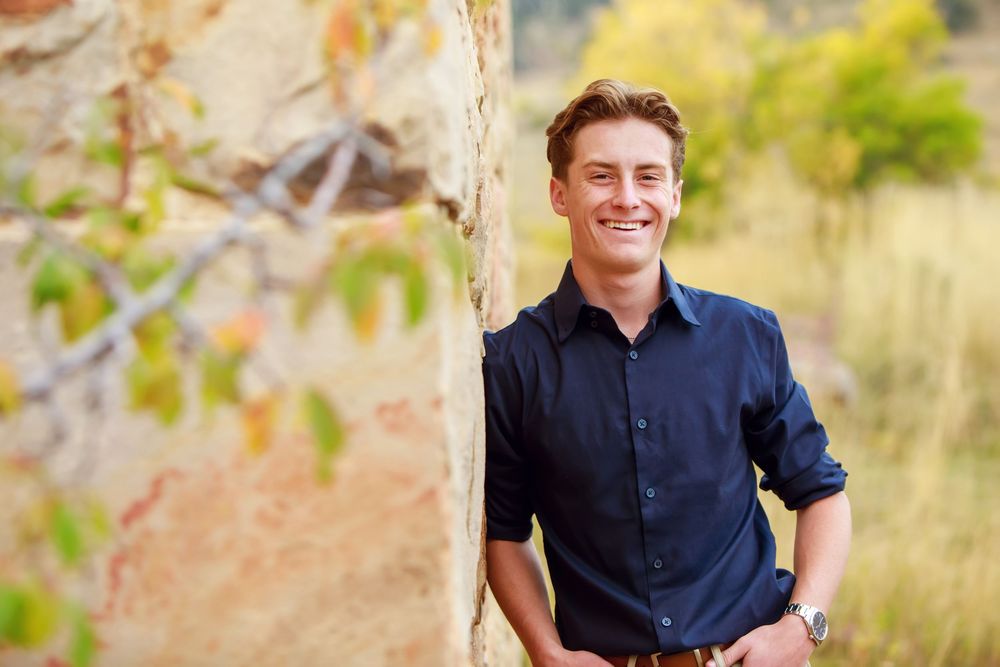A young person in a navy button-down shirt smiles outdoors with autumn foliage and natural scenery in the background.