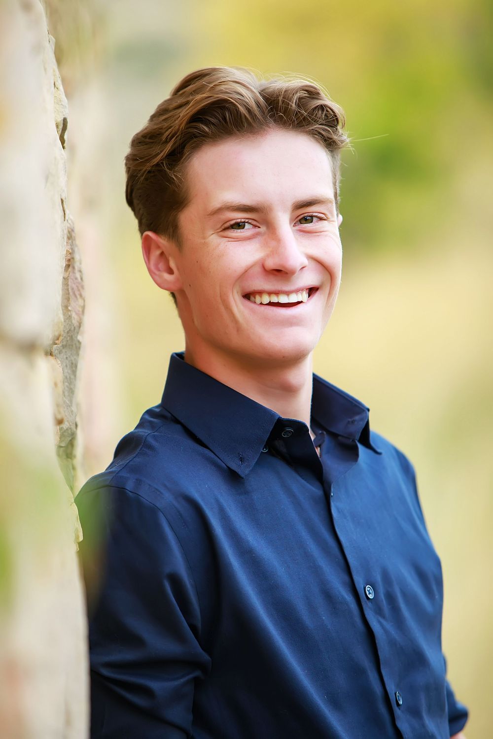 Professional portrait of a young person in a navy blue button-down shirt smiling against a blurred natural background.
