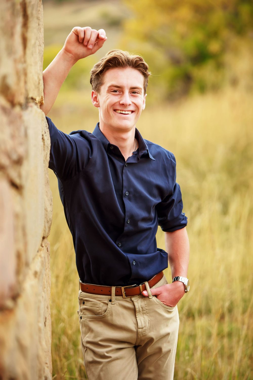 A person in a navy button-down shirt and khaki pants poses casually against a tree in a natural outdoor setting.