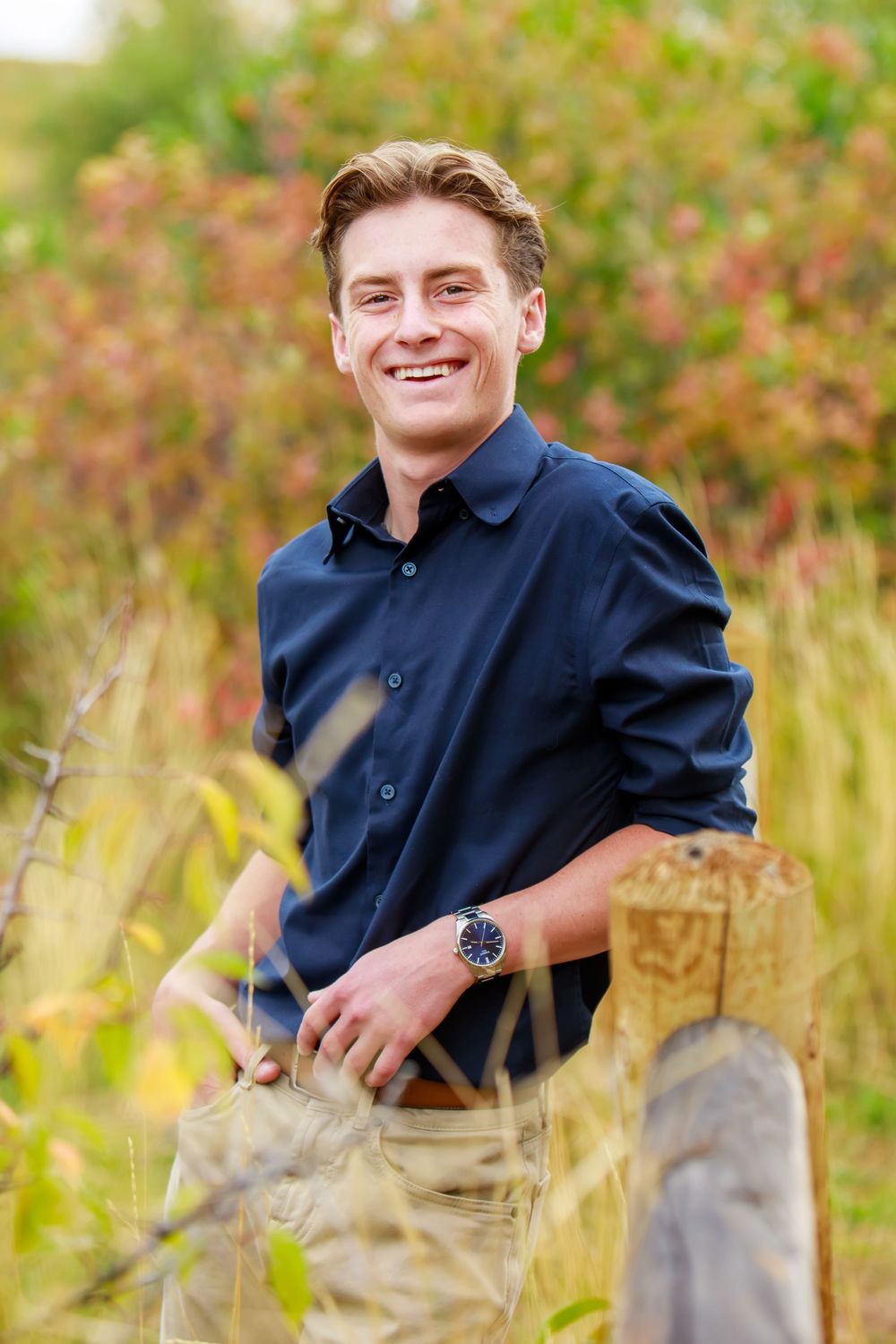 A person in a navy blue jacket sits outdoors among tall grass and autumn foliage on a sunny day.