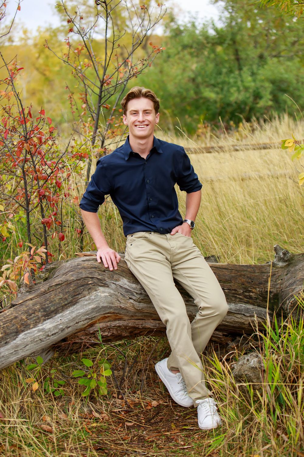 A person in a navy shirt and khaki pants sits casually on a fallen log in a natural autumn setting.
