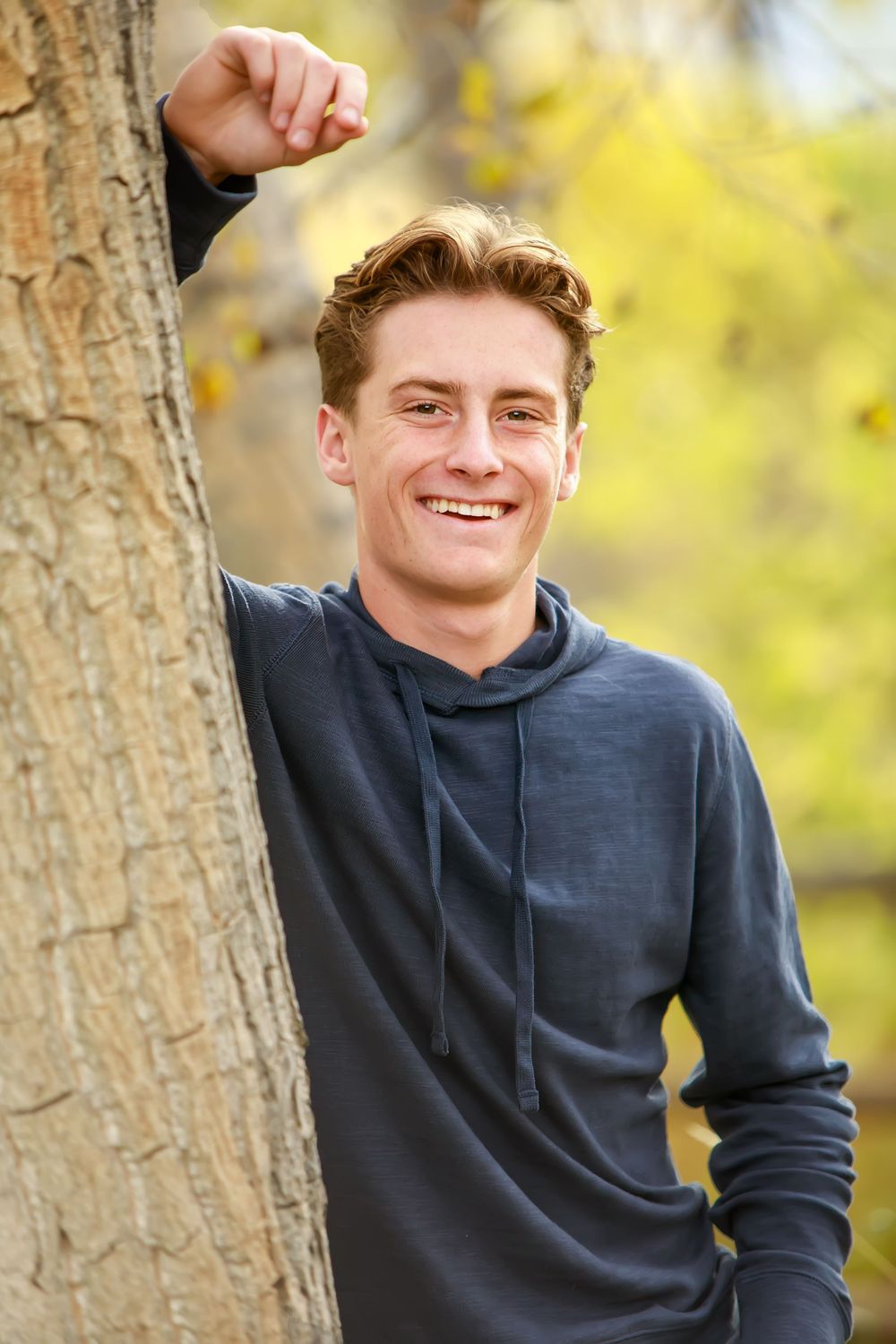 A person in a dark blue hoodie poses outdoors against a backdrop of autumn trees with golden foliage.