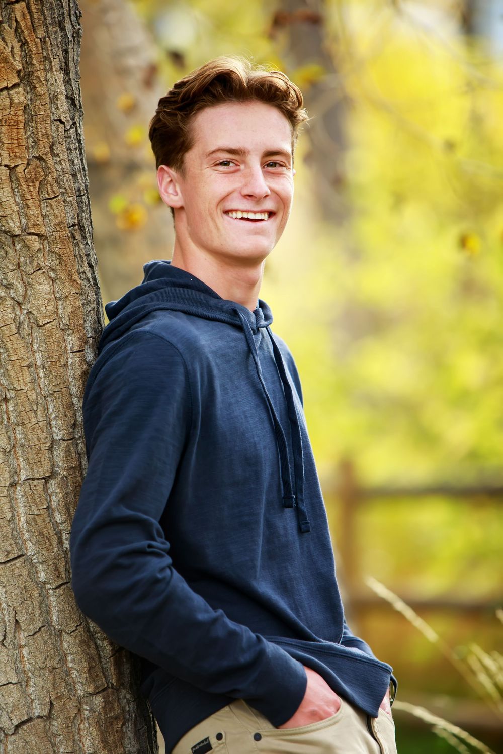 Young person in navy hoodie smiling while leaning against tree trunk with autumn foliage in background.