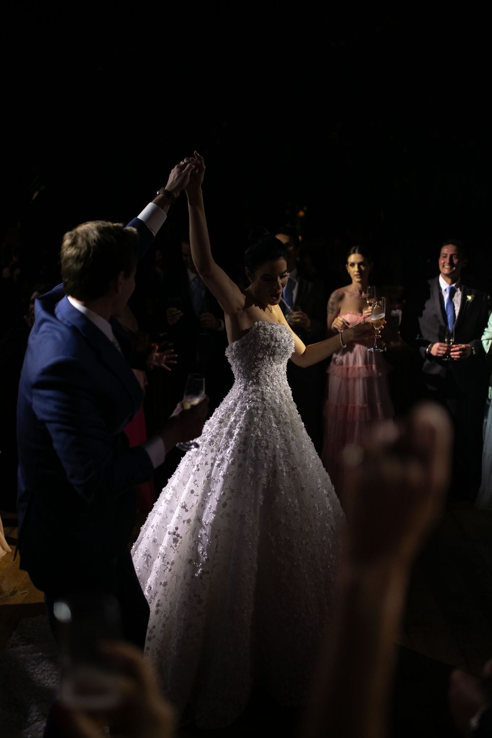 Wedding guests dance and celebrate on a dimly lit dance floor with a bride in a white lace gown.