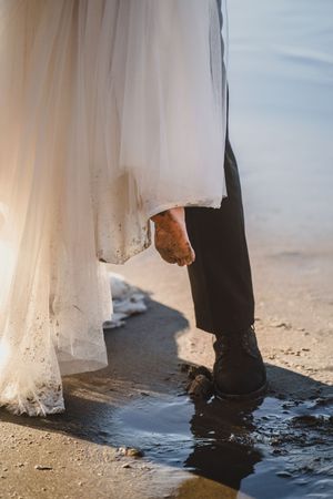 Close-up of bride and groom's feet standing in shallow water at beach during sunset wedding ceremony.