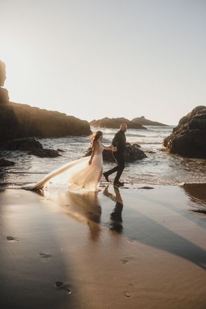 Couple walking along a misty beach shore with waves crashing against rocky outcrops in soft natural lighting.
