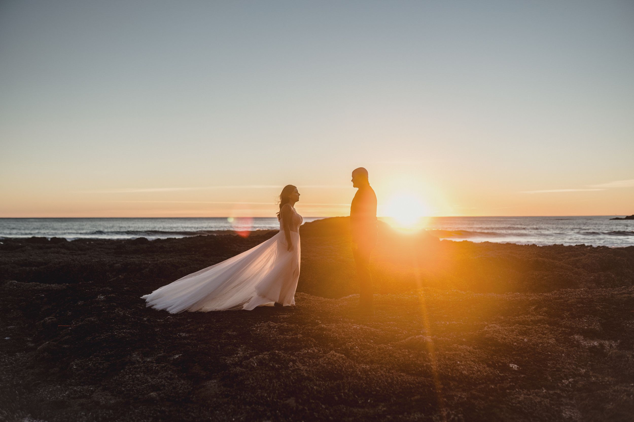 Two silhouettes sit on a cliff edge watching a golden sunset over the ocean horizon.