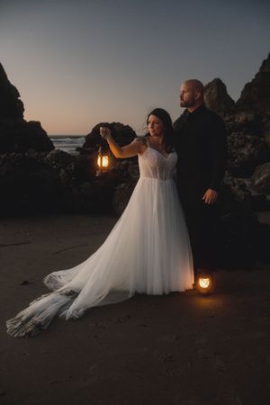 A couple poses with lanterns on a beach at dusk, surrounded by rocky cliffs and ocean waves.