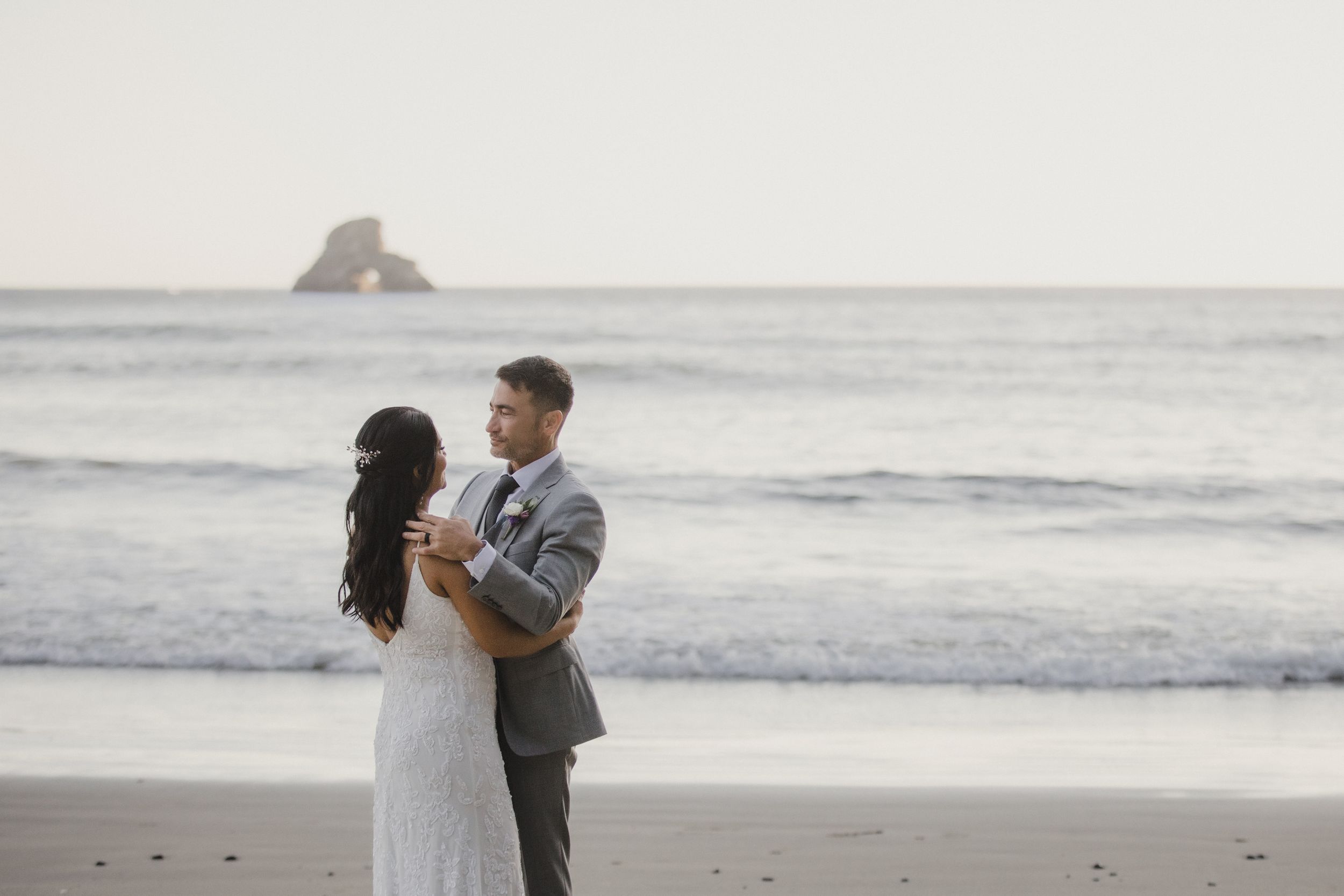 A couple in wedding attire embrace on a misty beach at dusk with waves and rocky coastline in the background.
