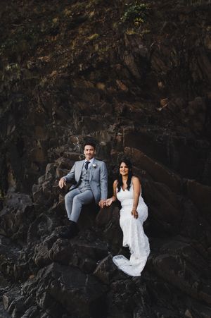 Newlywed couple sits together against dramatic dark rocky cliff face at sunset.
