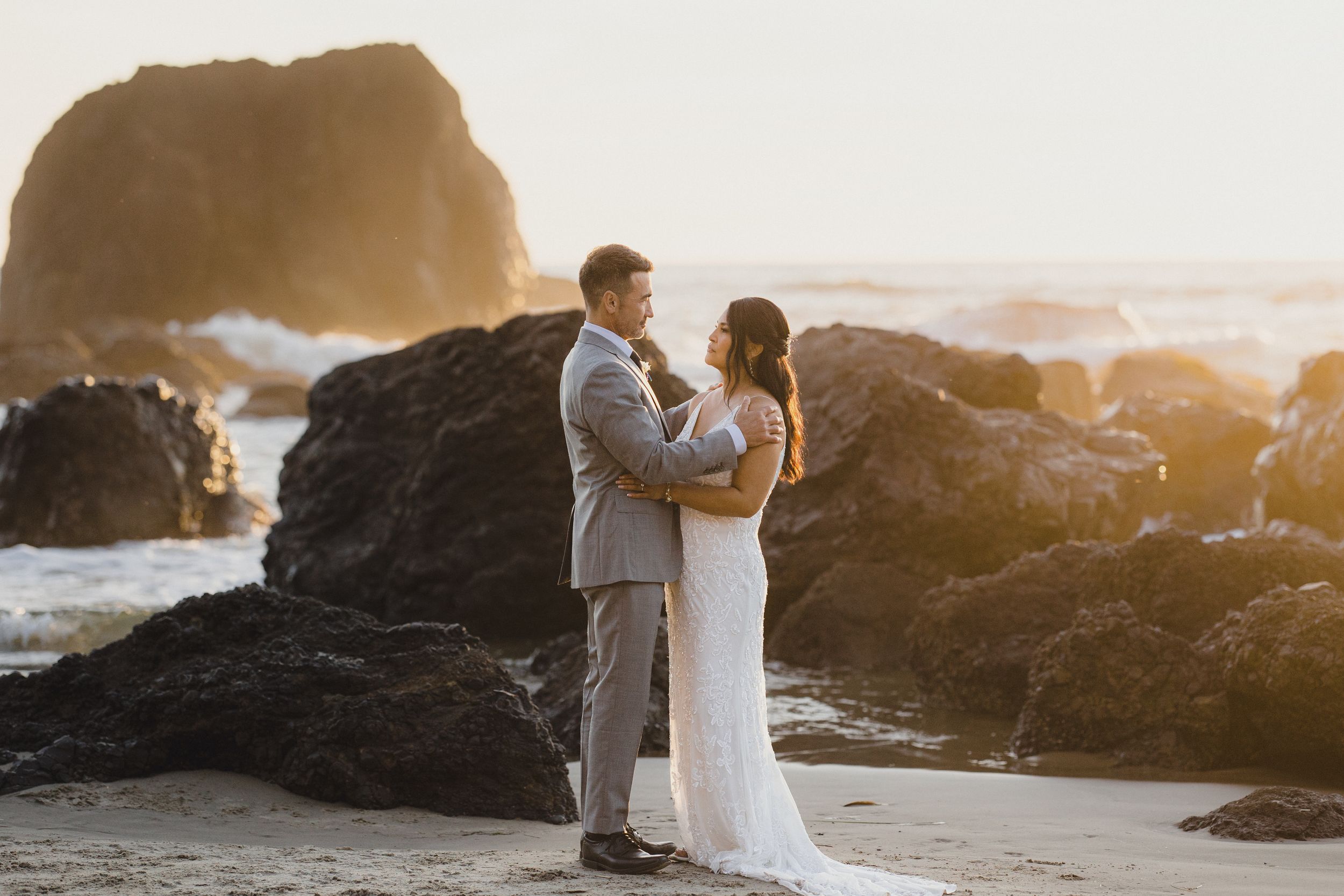 A couple shares an intimate moment at sunset on a rocky beach as golden light illuminates the coastal landscape.