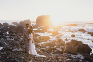 A couple in wedding attire poses on rocky coastal cliffs at sunset with ocean waves crashing in the background.
