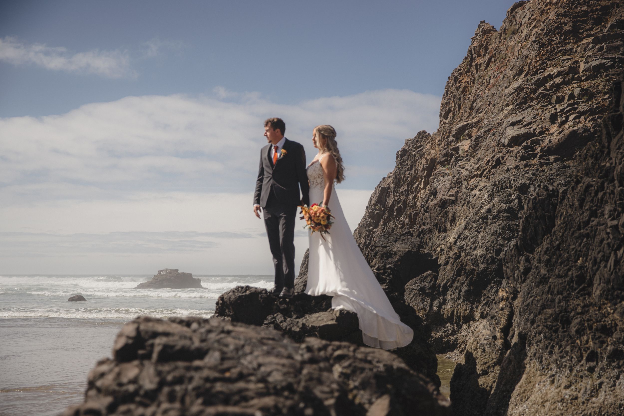 A couple in wedding attire poses together on rocky cliffs overlooking a dramatic ocean coastline at sunset.