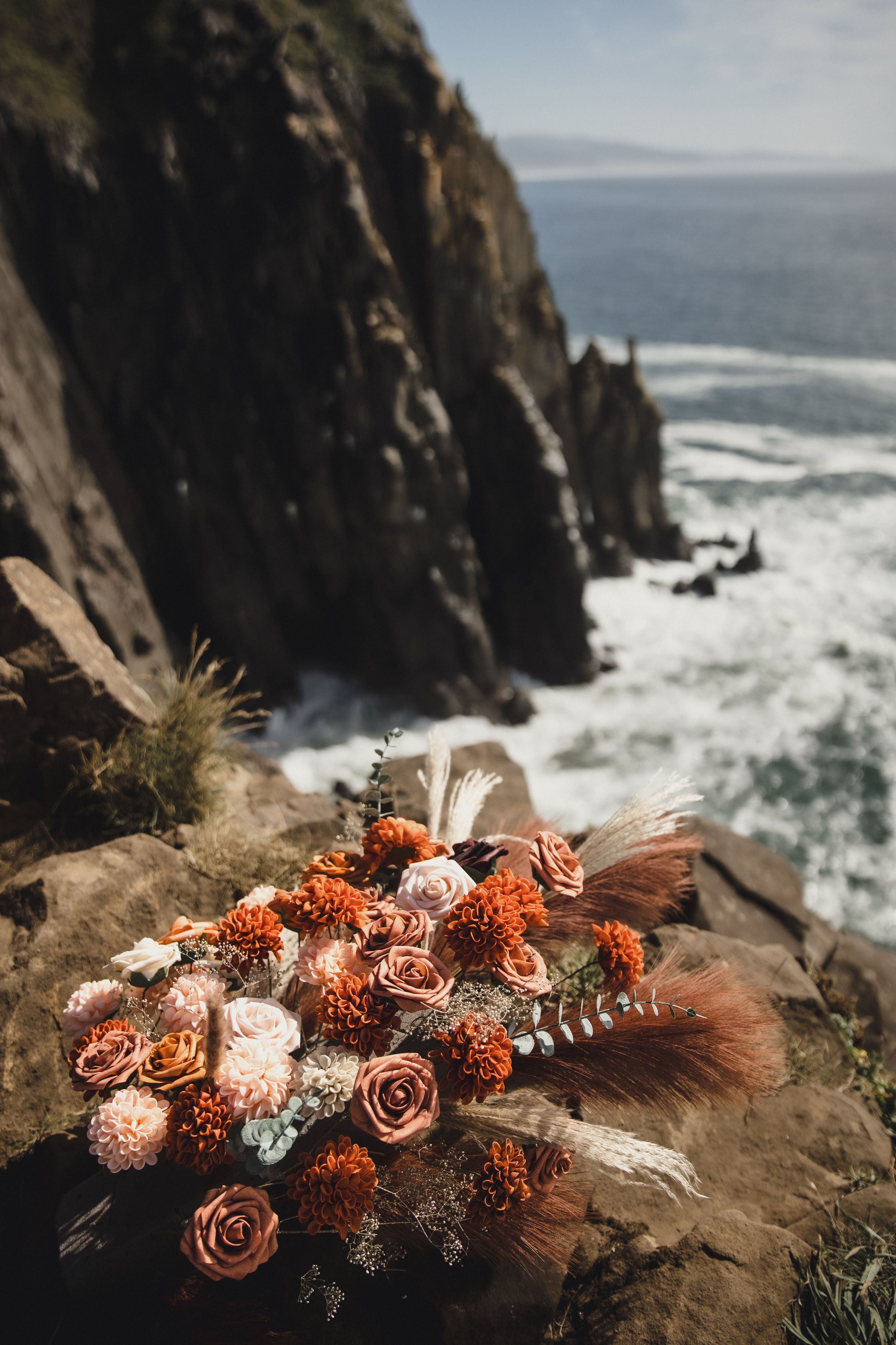 A rustic floral bouquet with orange and brown roses rests on a dramatic coastal cliff overlooking crashing ocean waves.