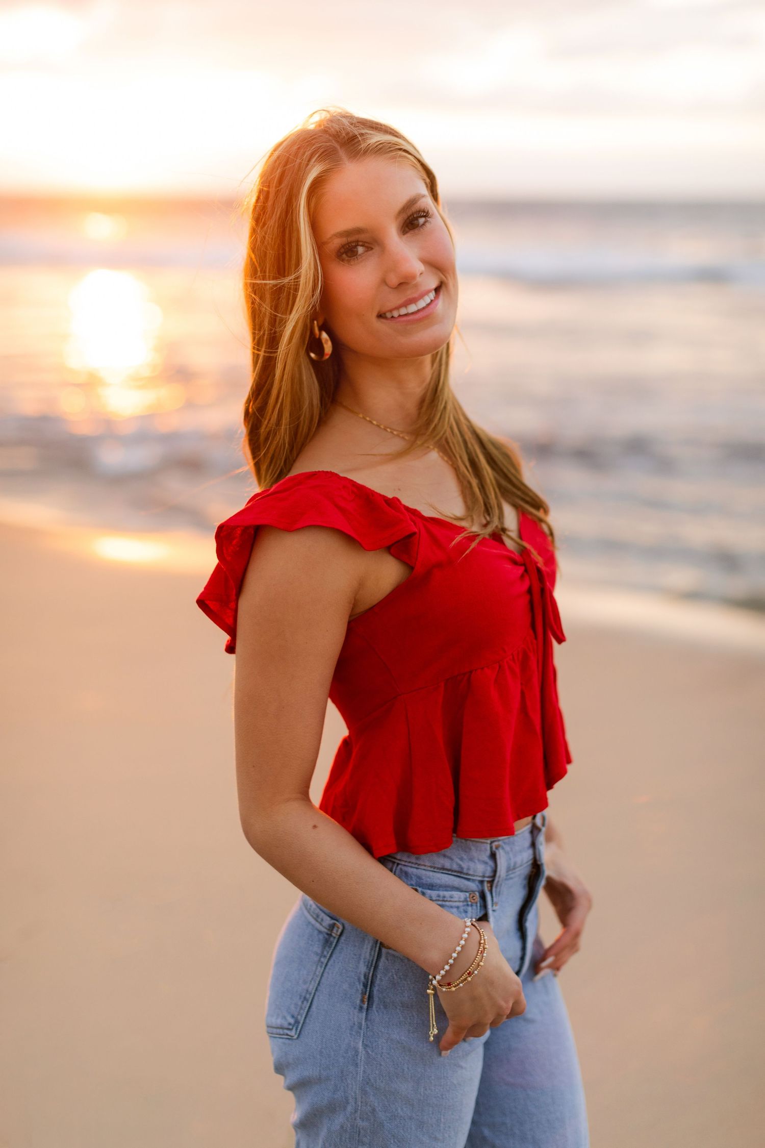 A figure in a red ruffled top and jeans poses on a beach at sunset with golden light illuminating the scene.