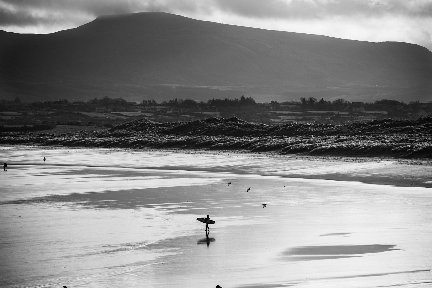 Dramatic black and white coastal landscape with surfers on a misty beach at sunset.