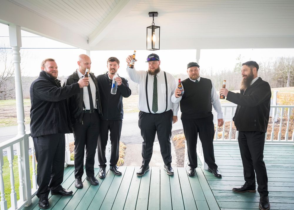 A panoramic photo of groomsmen in black suits posing playfully on a white porch railing.