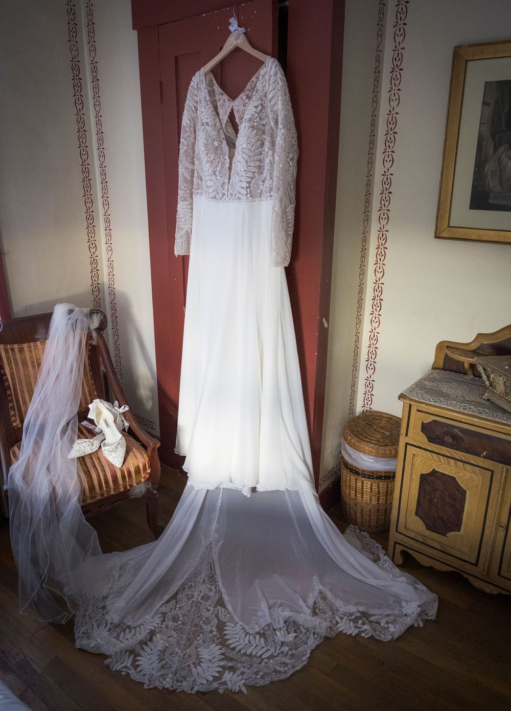 Elegant lace wedding dress with long train displayed in a vintage-style room with wooden furniture and floral wallpaper.
