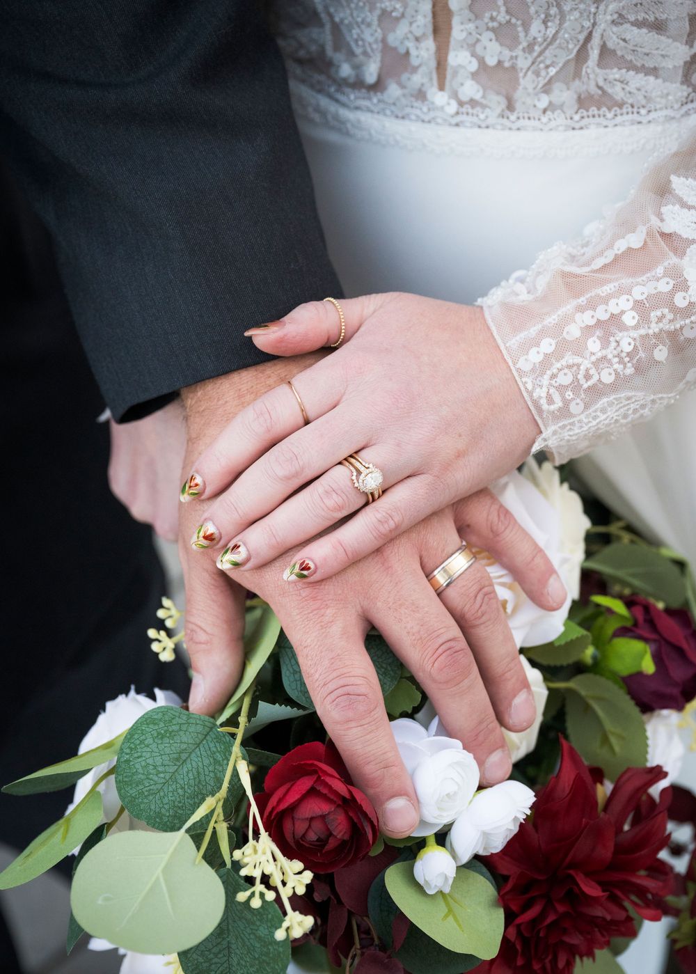 Wedding detail shot of hands with rings holding a deep red bouquet with greenery.