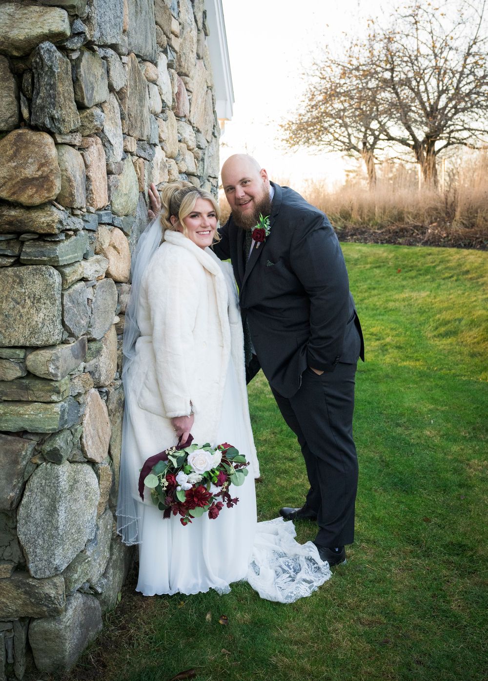 Wedding couple poses by stone wall outdoors during winter sunset.