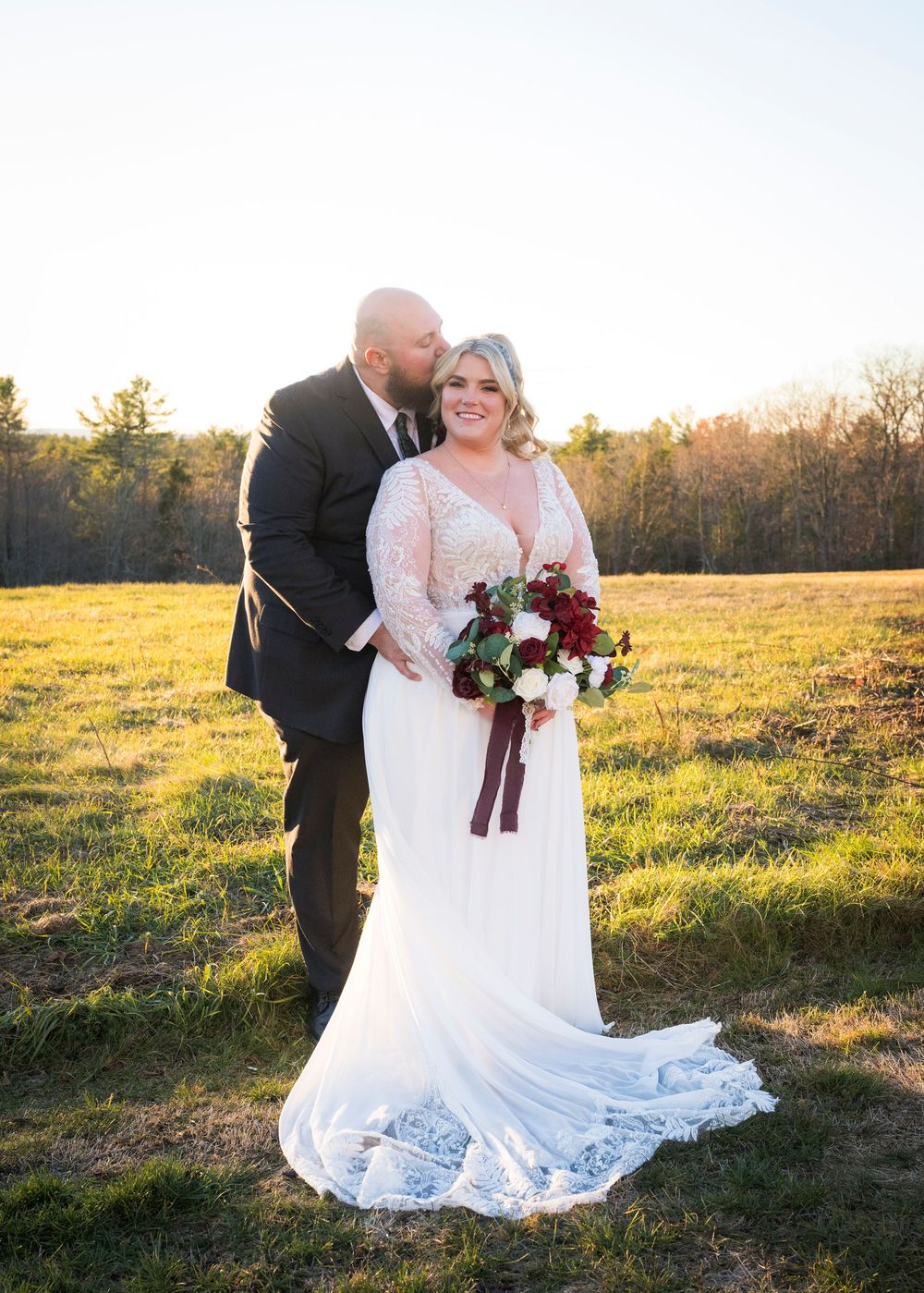 Newlyweds pose together in sunlit meadow with flowing white dress and burgundy bouquet.
