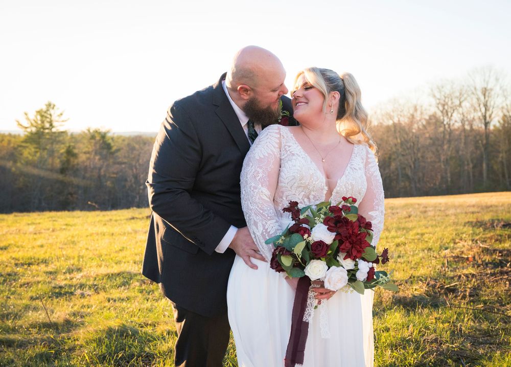 A couple shares a romantic moment in a sunlit field with burgundy floral bouquet at golden hour.