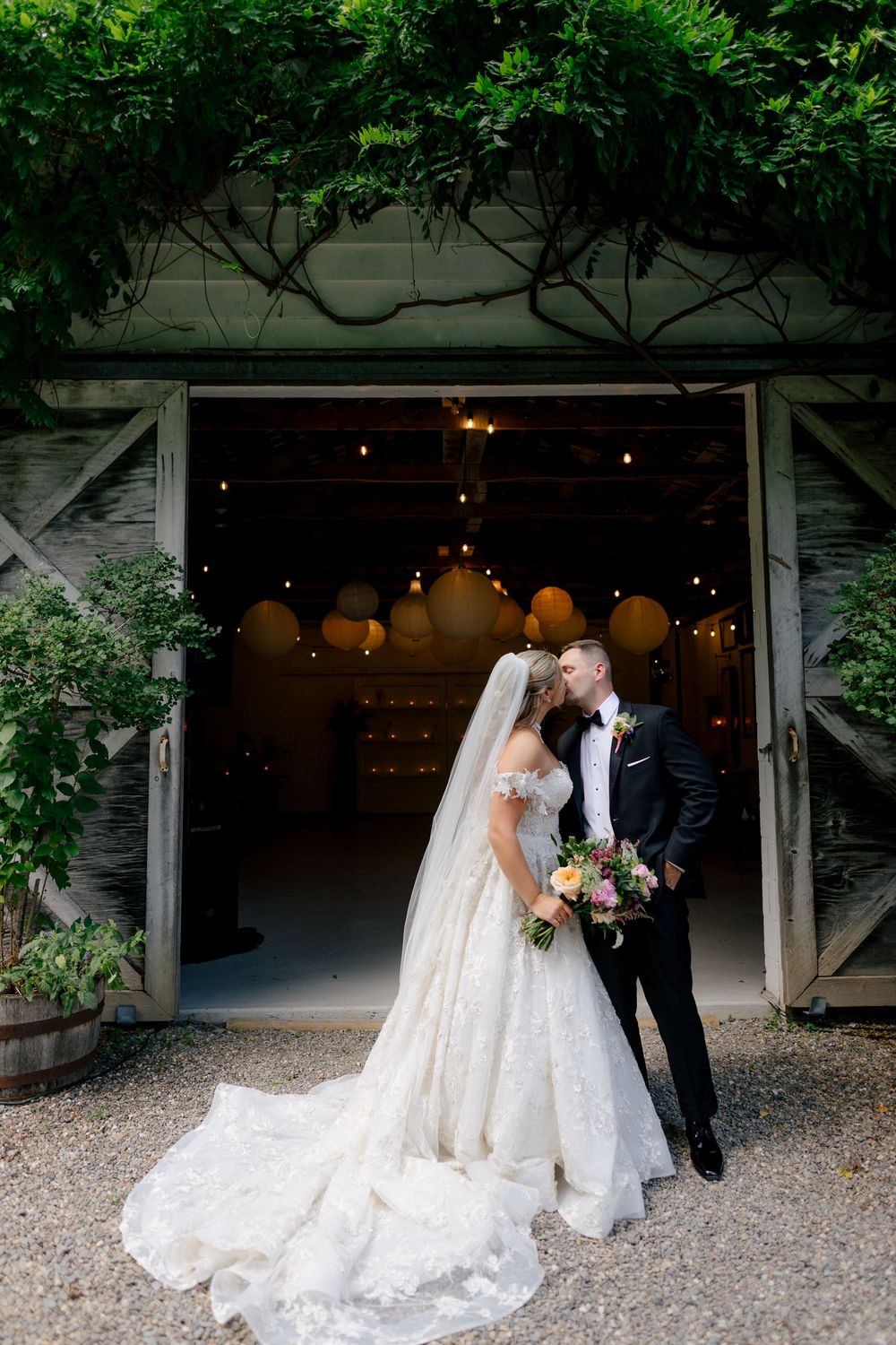 A wedding couple shares a kiss in front of a rustic barn entrance decorated with hanging lights.