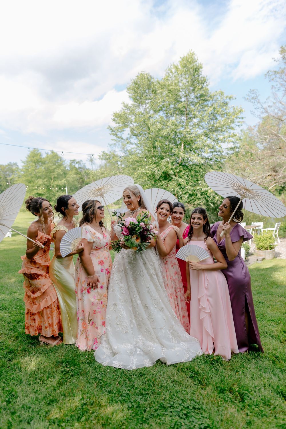 A wedding party in pastel dresses poses together on a grassy hillside with white tents in the background.