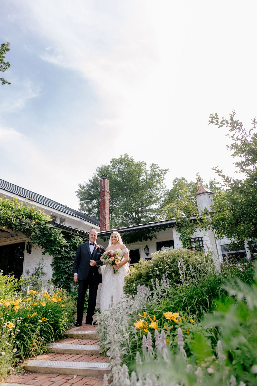 Wedding couple poses in a lush garden path with yellow flowers and greenery on a sunny summer day.