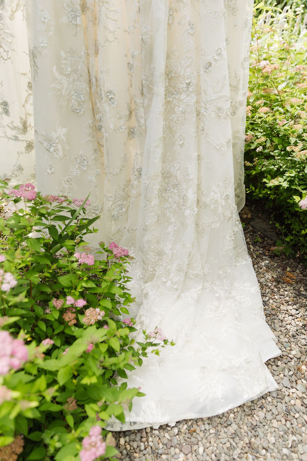 Delicate lace wedding dress hem trails along garden path beside blooming pink flowers and greenery.