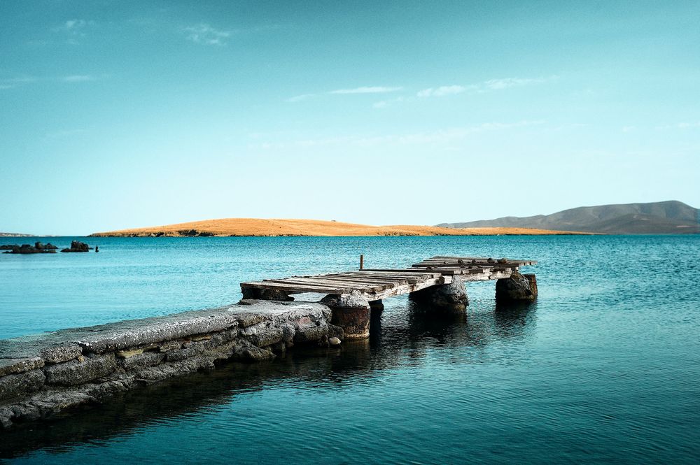 A weathered stone pier extends into turquoise waters with golden hills and blue sky in the background.