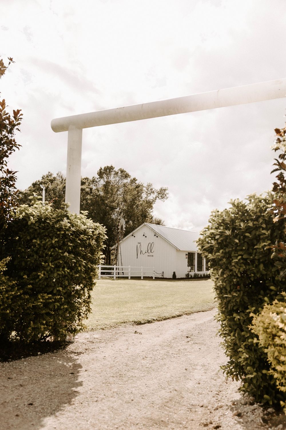 A white barn wedding venue with entrance path lined by bushes and trees.